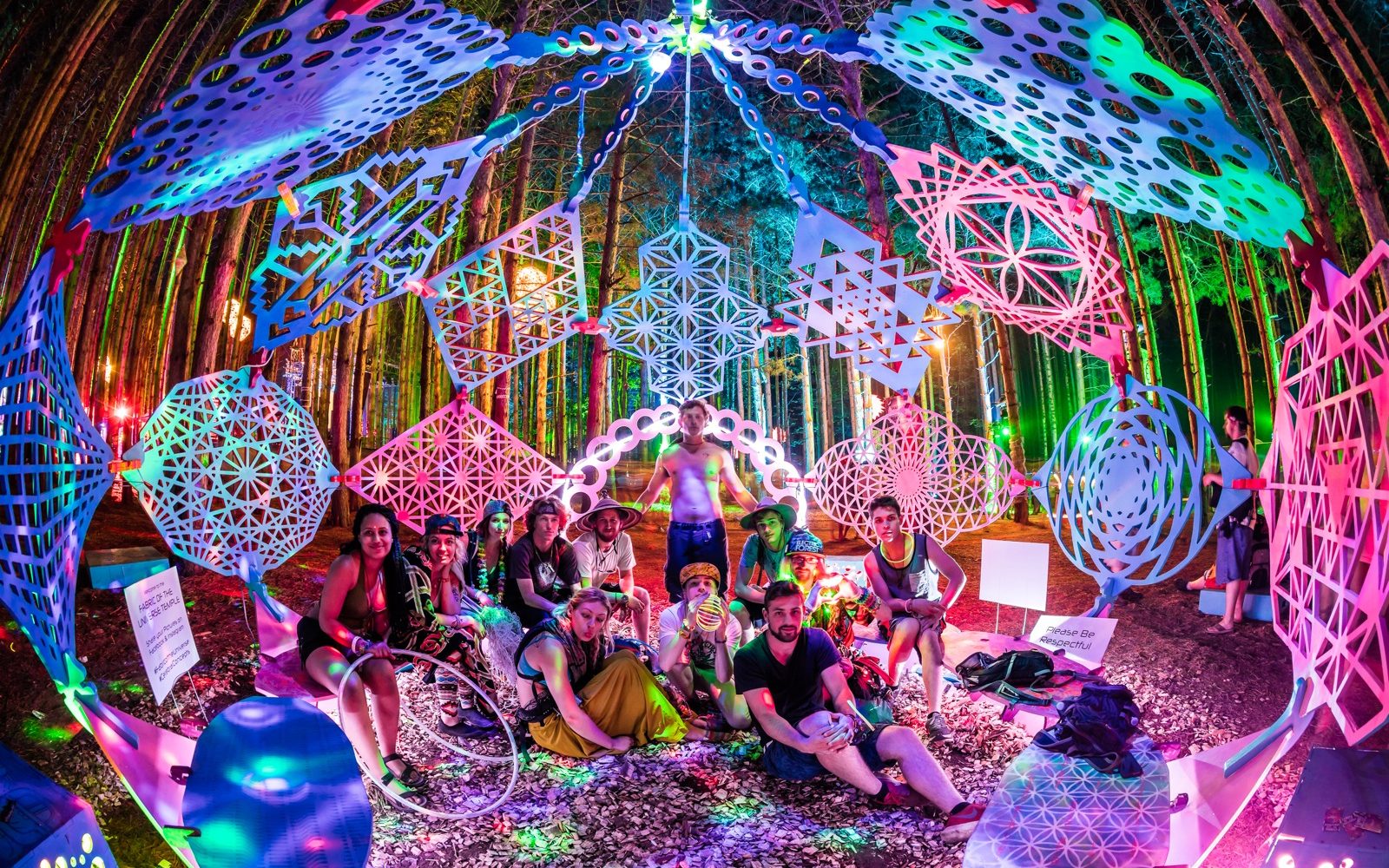 Group of twelve people sit in a orb made up of geometric shapes, in a forest, covered in colourful psychedelic light.