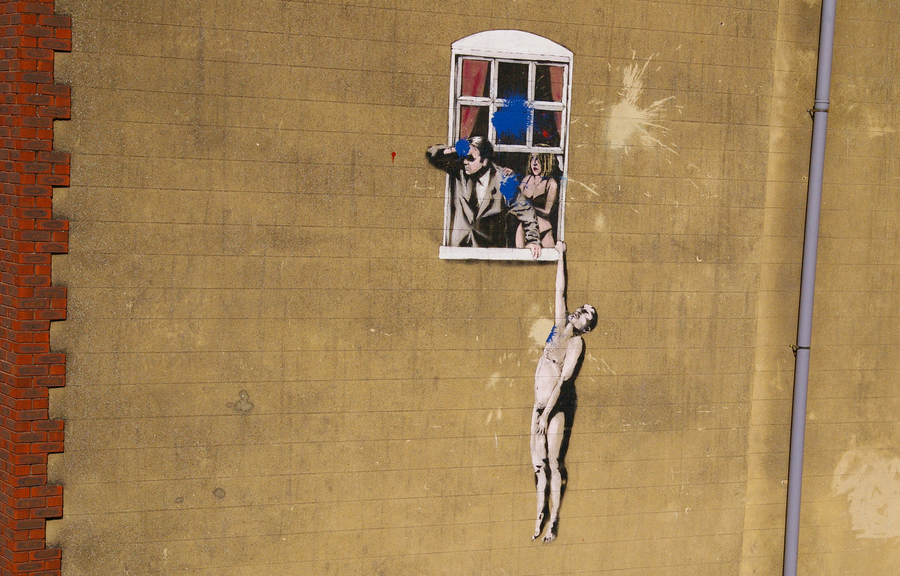 Banksy artwork on side the side of a building, man and woman stand in window, naked man hangs on window ledge