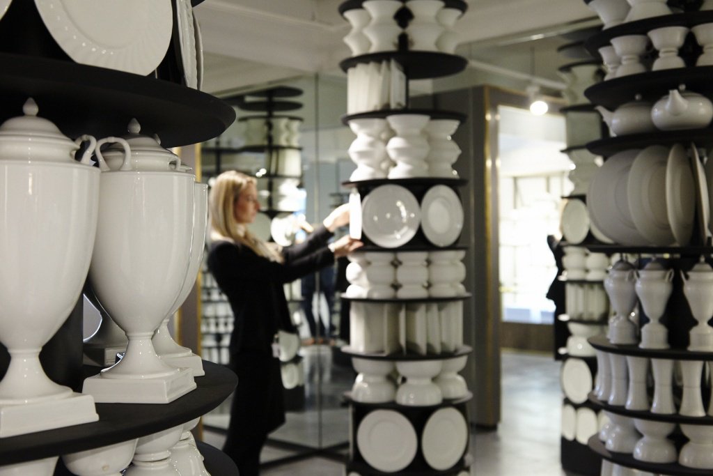 Various white, polished ceramic items displayed on shelves, wrapped around a number of columns. Female with long blond hair examines one of the items.