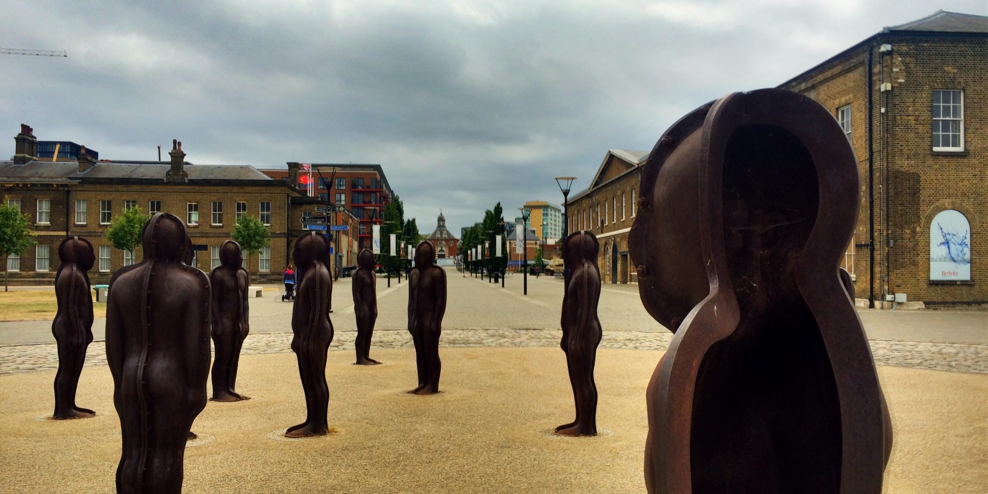 Eight figure shaped metal sculptures, stood on gravelled piazza with historic two storey buildings in background.