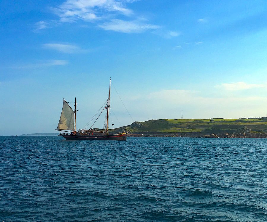 Old style, masted wooden sail boat on sea, with green landscape behind. A destination management plan for the Isles of Scilly