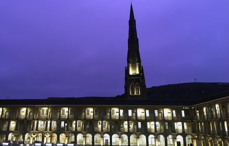 Courtyard within three storey building lit up at night, walkway on every storey around building supported by classical columns with railings around. Church spire in background