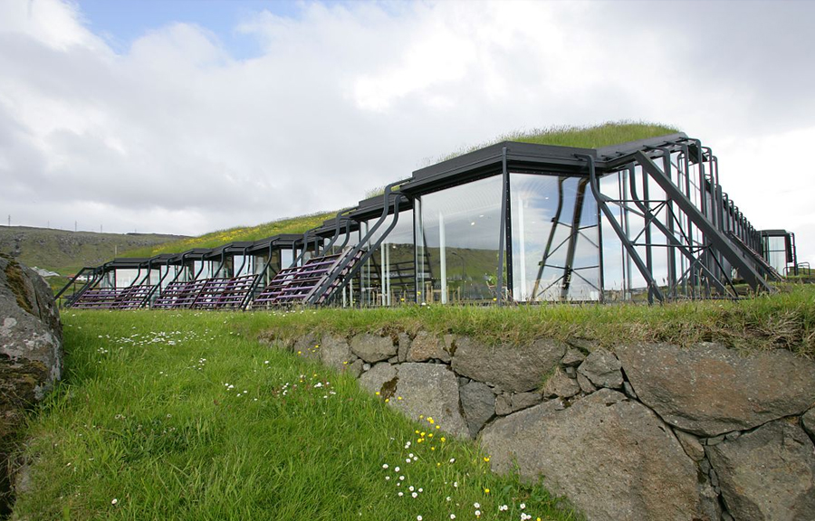 One storey glass panelled building, with metal supports, and organic grass roof. Surrounded by grass and stone.