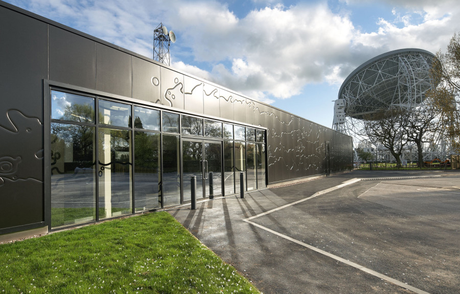 Single storey building with flat roof, covered in ornate black cladding, with glass entrance. Large white telescopic dish to the background - Jodrell Bank, University of Manchester