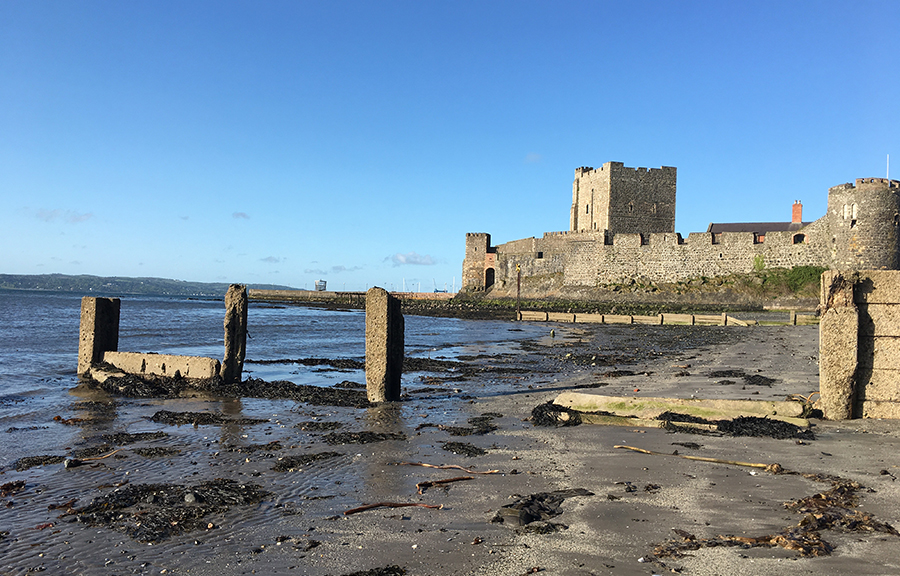 Carrickfergus Castle, a walled castle, sits on a beach, tide partly out, seaweed visible on sand.