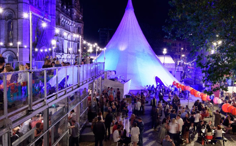 Crowded square in the evening. Large white teepee tent in the background, two storey hospitality area to the left, benched seating the right. Area decorated with red Chinese lanterns and festive lighting at MIF Festival Square.